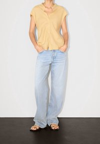Camicia gialla chiara a maniche corte con bottoni, jeans blu a gamba larga e sandali neri flat, in piedi su una superficie nera con uno sfondo bianco.