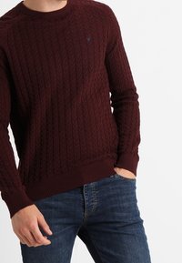 Pull en tricot bordeaux avec un motif torsadé, col rond et poignets côtelés, associé à un jean en denim foncé.