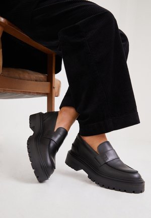 Person iført sorte chunky læderloafers med tykke såler og sorte vidde kordbuksbukser, der sidder på en brun stol.