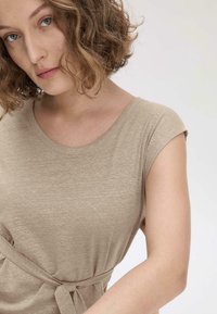 Beige Kurzarmoberteil aus weichem Material mit Rundhalsausschnitt und gebundenem Bund für eine figurbetonte Silhouette. Subtile Textur verleiht Tiefe.