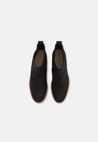 Bottines en daim noir avec une texture lisse, des panneaux latéraux élastiques et une semelle en caoutchouc brun, arborant un design minimaliste et sans éléments matériels visibles.