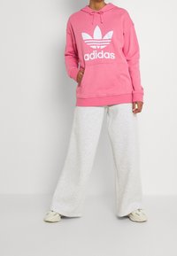 Rosa hoodie med vit Adidas-logga och tre ränder; ihopparad med lösa ljusgråa sweatsbyxor och beige sneakers. Mjuk tygtextur synlig.