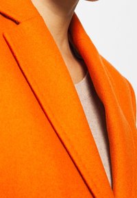 Lyseorange uldblanding frakke med en indsnitkrave og blød tekstur, lagdelt over en lysebeige sweater. Nærbillede af stof og farve.