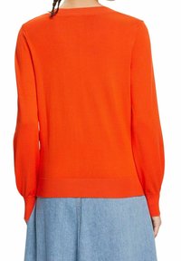 Femme portant un pull à manches longues orange vif et une jupe en jean bleu clair, vue de dos.