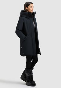 Sort vandtæt parka med hætte, med lige snit, lynlås foran og sidelommer. Kombineret med isolerede sorte snesko.