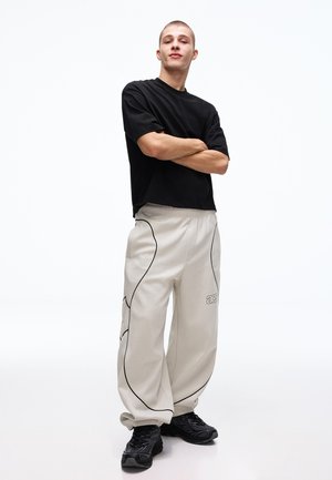 Ung mand står selvsikkert med armene krydset, iført en sort T-shirt, løse beige bukser med sorte kanter og sorte sneakers på en hvid baggrund.