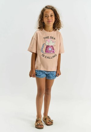 Vaaleanpunainen puuvillainen t-paita, jossa vintage-tyylinen puhelin ja teksti "THE SEA IS CALLING." Yhdistetty valkoisilla pilkuilla koristeltuihin farkkushortseihin.