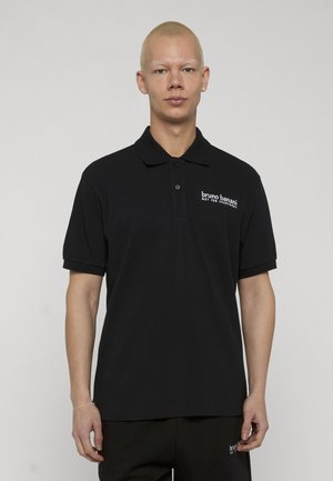 Junge Person mit kurzen blonden Haaren, die ein schwarzes Poloshirt mit dem Schriftzug "bruno banani NOT FOR EVERYBODY" auf der Brust trägt.