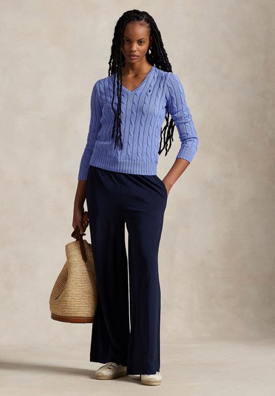 Kobieta z długimi warkoczami, ubrana w jasnoniebieski sweter z warkoczowym splotem, granatowe szerokie spodnie i białe buty, trzymająca dużą plecioną torbę na ramię.