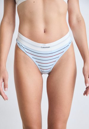 Woman wearing light blue striped Calvin Klein high-cut underwear and white sports bra, standing against a plain background.