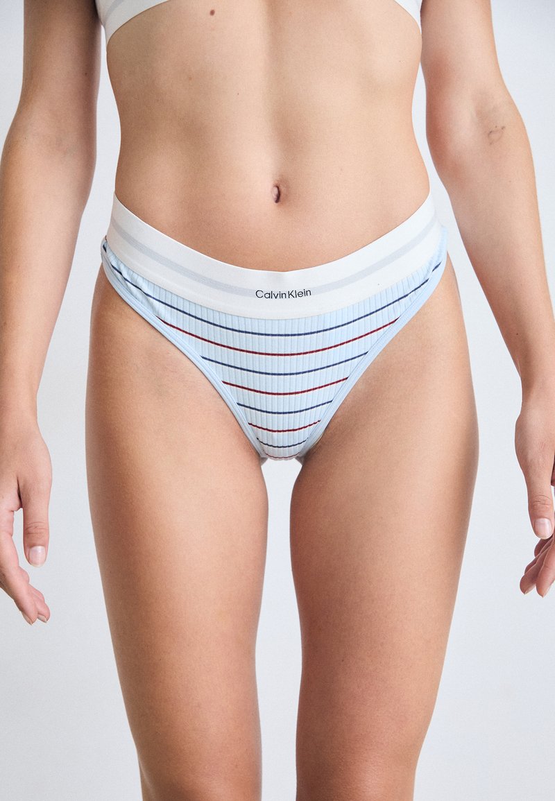 Žena u svijetloplavim prugastim Calvin Klein gaćicama visokog struka i bijelom sportskom grudnjaku, stoji protiv jednostavne pozadine.