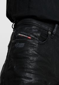 Svarta denimjeans med en sliten textur, med en synlig framficka och en röd märkesetikett sydd p å midjebandet.