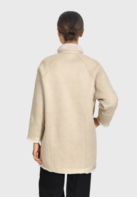 Un abrigo beige de corte oversized con cuello, hecho de material suave. Presenta una superficie texturizada, mangas largas y forro de forro polar contrastante en el dobladillo.