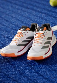 Witte sportief schoenen met oranje en zwarte accenten, een bovenwerk van mesh, contrasterende strepen en gevoerde zolen, geplaatst op een blauwe textuur oppervlak.