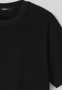 T-shirt noir ample avec une texture côtelée, col rond et manches courtes. Le tissu semble doux ; l'étiquette est visible au niveau du col.