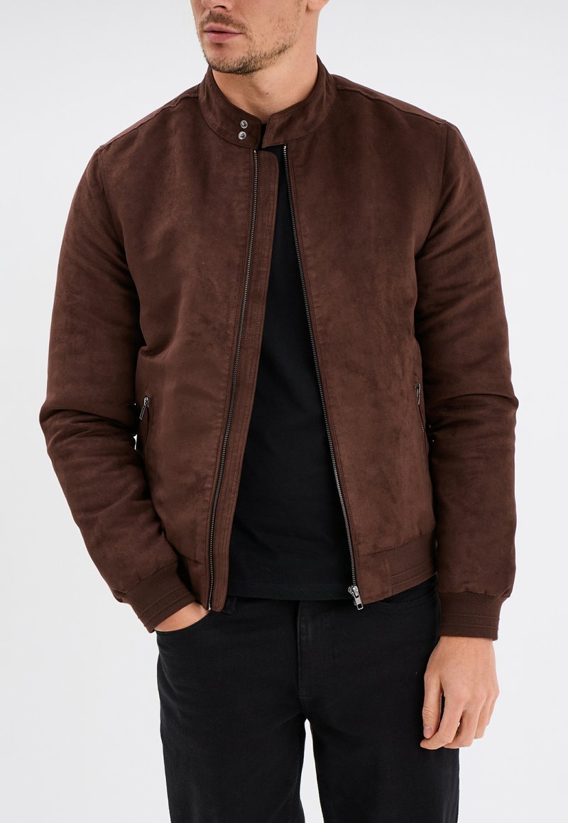 Bruine leren bomberjack van suede met een zachte textuur, korte kraag, ritssluiting en zijzakken, gedragen met een zwart shirt en jeans.