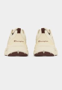 Ζεύγος υποδημάτων Champion σε εκρού χρώμα με καφέ ταμπελάκια και λογότυπα στη φτέρνα, φωτογραφημένα από πίσω σε απλό λευκό φόντο.