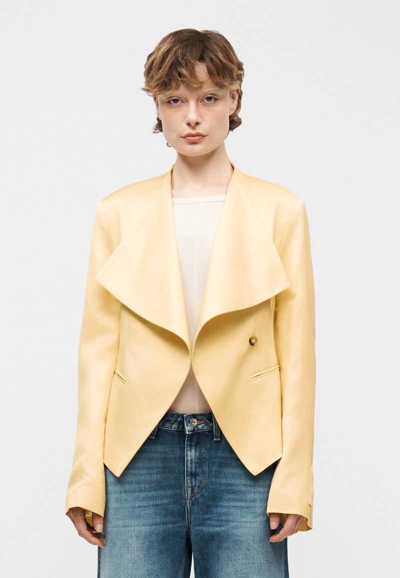 Άτομο με κοντά μαλλιά φοράει ένα ανοιχτό κίτρινο ασύμμετρο σακάκι πάνω από λευκή μπλούζα και μπλε τζιν, σε απλό λευκό φόντο.