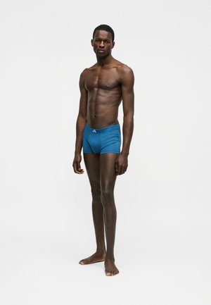 Αθλητικός άνδρας που στέκεται ξυπόλυτος φορώντας σφιχτό μπλε μποξεράκι, με φόντο απλό λευκό.