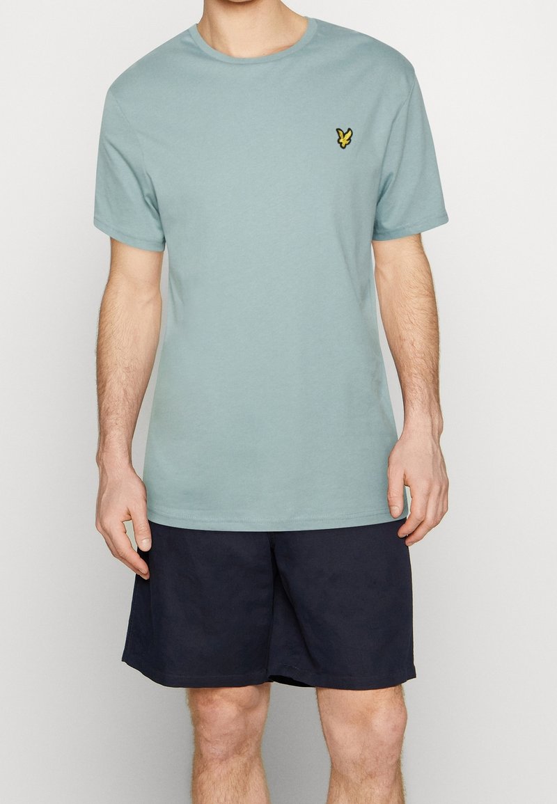 Uomo che indossa una maglietta a maniche corte di colore azzurro chiaro con un piccolo logo giallo sul petto e pantaloni corti blu scuro.