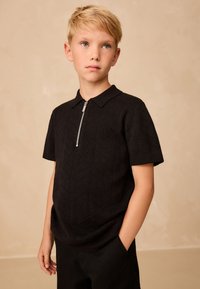 Czarna koszulka polo z krótkim rękawem, z zamkiem przy kołnierzu i geometrycznym fakturowanym wzorem, wykonana z miękkiej tkaniny, zestawiona z ciemnymi spodniami.