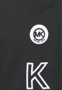Svart tyg med ett framträdande vitt broderat "K" och en cirkulär logotagg med "MK" och "MICHAEL KORS" i svart på vit bakgrund.
