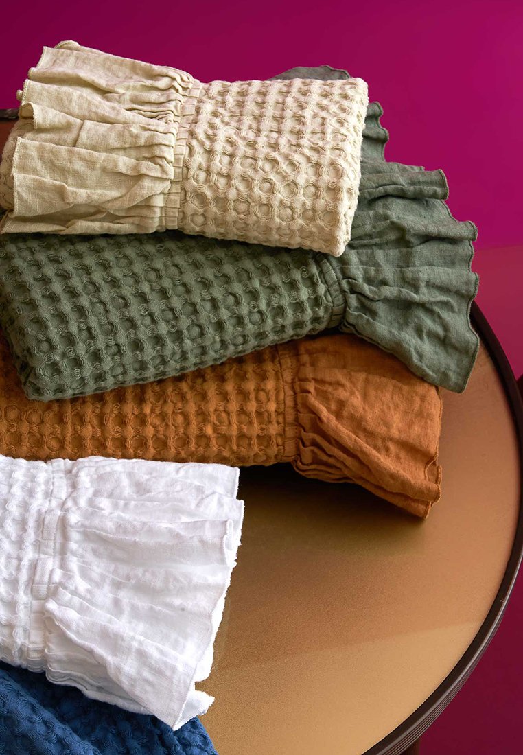 Stapel gefalteter, strukturierter Stoffstücke in Creme, Grün, Rost, Weiß und Blau, auf einem runden braunen Tisch vor einem magentafarbenen Hintergrund.