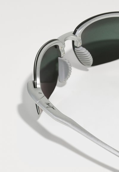 Lunettes de soleil argentées avec des verres verts, présentant un design élégant, des coussinets de nez texturés et un détail signature sur la branche.