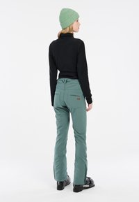 Grüne Skihosen im Slim-Fit-Stil mit Reißverschluss an den Knöcheln, kombiniert mit einem schwarzen, gerippten Langarmshirt und einer hellgrünen Strickmütze.