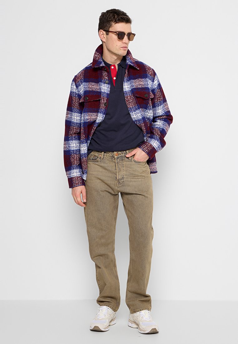 Camicia a quadri rossa, blu e bianca con due tasche sul petto, indossata sopra una polo blu navy, abbinata a jeans beige e sneakers bianche.