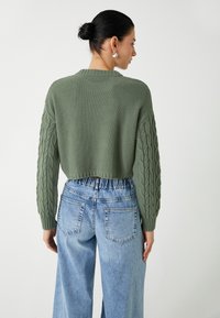 Grüner Strickpullover mit strukturierten Kabelärmeln, kombiniert mit hoch taillierten hellblauen Denim-Jeans. Einfache runde Ohrringe sind sichtbar.