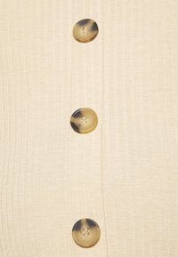 Trois boutons ronds en écaille sur un tissu côtelé couleur crème avec des détails de couture verticale.