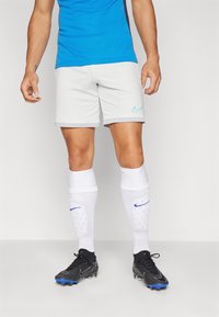 Shorts de sport Nike gris clair, design ajusté, avec un logo turquoise sur le côté gauche. Associés à des chaussettes blanches jusqu'au genou et des chaussures de football noires.