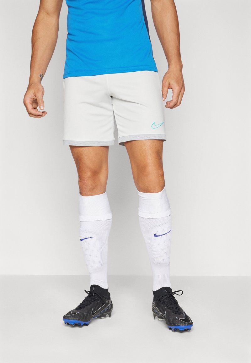 Shorts de sport Nike gris clair, design ajusté, avec un logo turquoise sur le côté gauche. Associés à des chaussettes blanches jusqu'au genou et des chaussures de football noires.