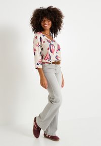 Haut beige avec des motifs de cœurs colorés, manches trois-quarts ; jean évasé gris clair ; baskets bordeaux avec des rayures roses ; ceinture marron.