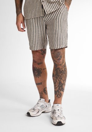 Striped shorts in beige and navy fabric, featuring a relaxed fit and side pockets, paired with white athletic sneakers. Tattoos visible on legs.