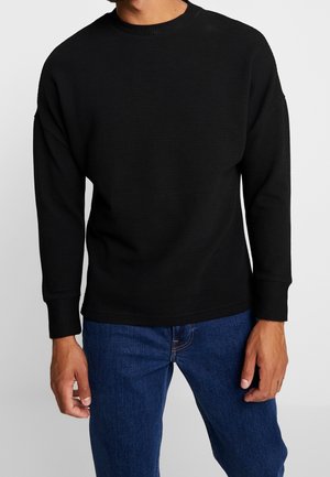 Mand iført en sort ribstrikket langærmet sweater og blå cowboybukser, stående mod en ensfarvet hvid baggrund.