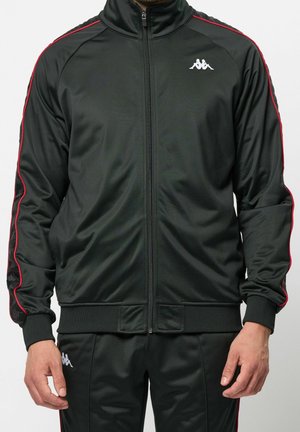 Mand iført sort sportjakke med lynlås og bukser med røde striber og hvidt logo på bryst og lår, hænder afslappet ned langs siden.