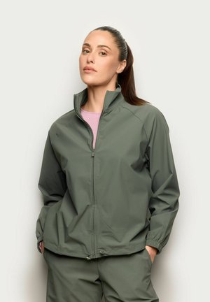 Frau mit braunen Haaren im Pferdeschwanz, die eine grüne Jacke mit Reißverschluss und passende Hose trägt, mit Händen in den Taschen vor einem schlichten Hintergrund.