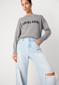 Suéter gris de punto con el texto "calvin klein", combinado con jeans rasgados de color azul claro. El atuendo presenta un corte relajado y un escote redondo.