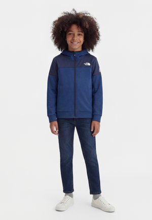 Παιδί με σγουρά μαλλιά που φοράει μπλε φούτερ με φερμουάρ, σκούρο τζιν, γκρι κάλτσες και λευκά αθλητικά παπούτσια, στέκεται μπροστά σε ένα απλό λευκό φόντο.