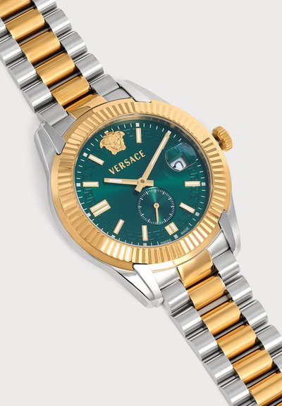 Montre-bracelet bicolore argent et or avec un cadran vert, des index et aiguilles dorés, logo Versace, et une loupe pour la date à trois heures.
