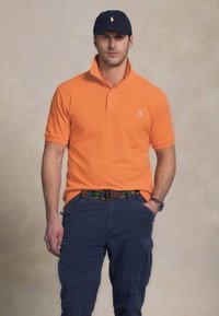 Tricou polo portocaliu cu guler, mic logo pe piept, asortat cu pantaloni cargo bleumarin și o șapcă bleumarin cu logo. Cureaua are accente verzi și galbene.