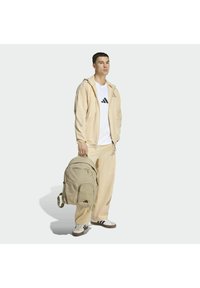 Chaqueta ligera beige sobre una camiseta blanca con un logo negro, combinada con pantalones beige y una mochila texturizada color kaki con dos bolsillos frontales.