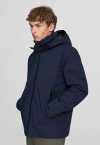 Giovane uomo in una giacca con cappuccio blu navy, munita di tasche con zip, indossa un scaldacollo abbinato e si trova davanti a uno sfondo semplice.