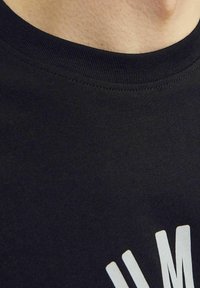 Juodas medvilnės marškinėliai su apvaliu kaklu ir raukšlėtu apykaklę, priekyje su baltu tekstu, lygi tekstūra ir prigludusi forma.