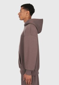 Giovane con capelli ricci corti, indossa un pantalone e una felpa oversize marroni, in profilo su uno sfondo semplice.