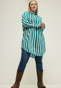 Camisa de botones a rayas verde y blanca con mangas largas, dobladillo asimétrico, combinada con jeans azules y botas de vaquera marrones.