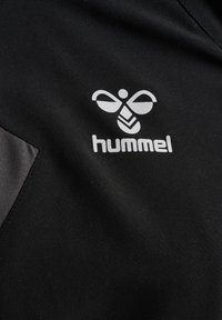 Sort stof med hvidt styliseret bi-logo og "hummel" tekst, del af et sportsbeklædningsplagg.