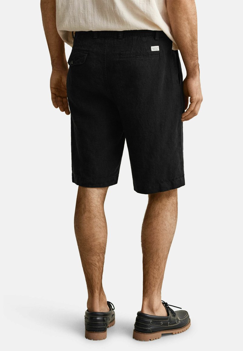 Hombre con pantalones cortos negros hasta la rodilla y zapatos casuales negros, de pie contra un fondo claro y liso.